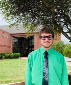 A smiling individual in a green shirt and black tie standing in front of a brick building at Howard Payne University with the sign "school of music". | HPU