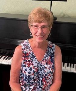 A smiling woman with short hair wearing glasses and a floral top sitting in front of a piano at Howard Payne University. | HPU