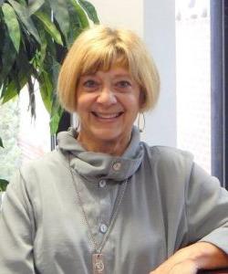 Smiling woman with short blonde hair, wearing a Howard Payne University gray outfit and necklace, seated indoors. | HPU